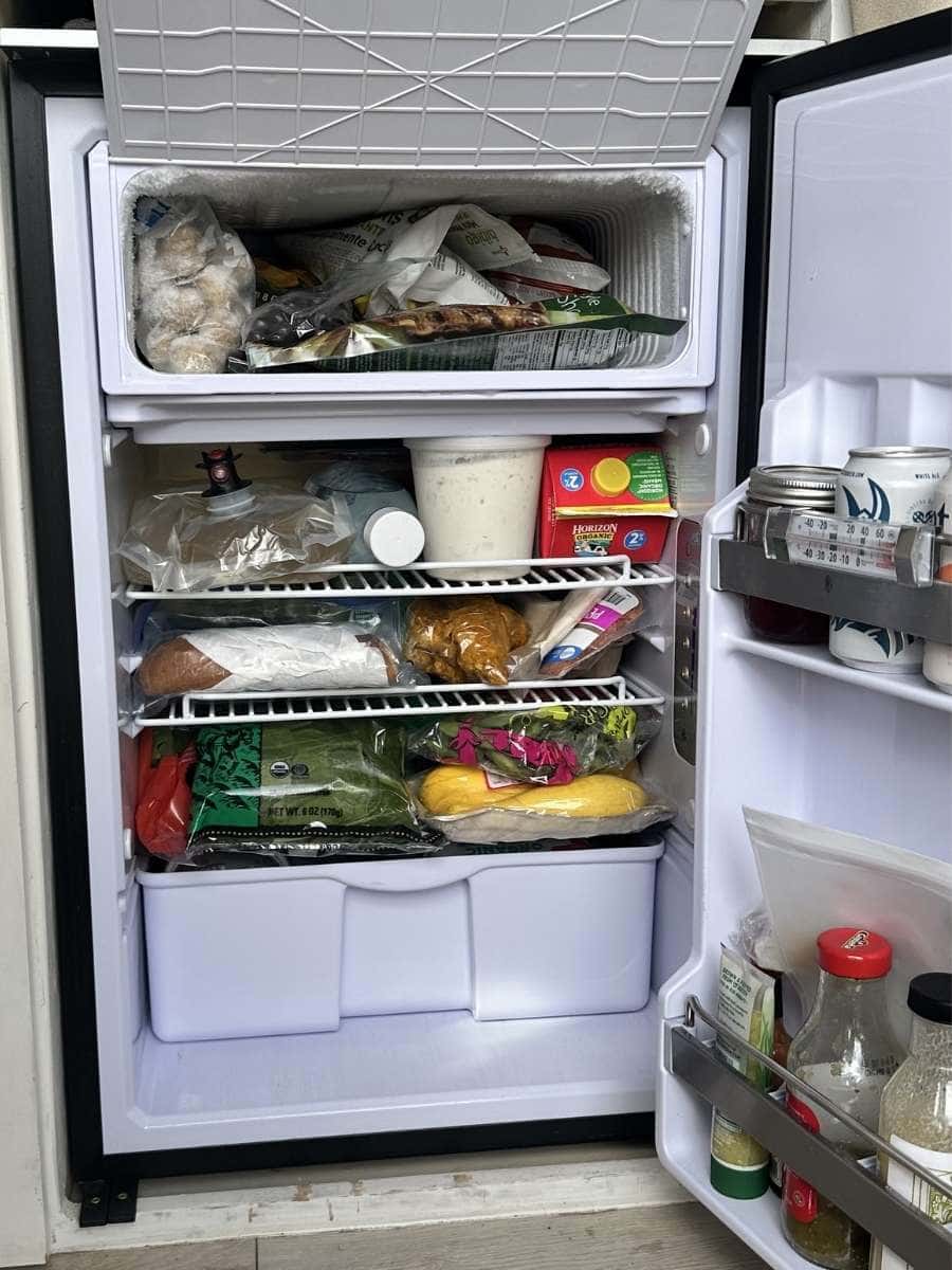 Isotherm refrigerator review Interior fridge view featured in Isotherm refrigerator reviews showing a compact freezer compartment filled with frozen food above multiple wire shelves stocked with groceries. Items include leafy greens, squash, packaged foods, jars, and condiments in the door, illustrating how much can fit inside a small fridge for everyday use.