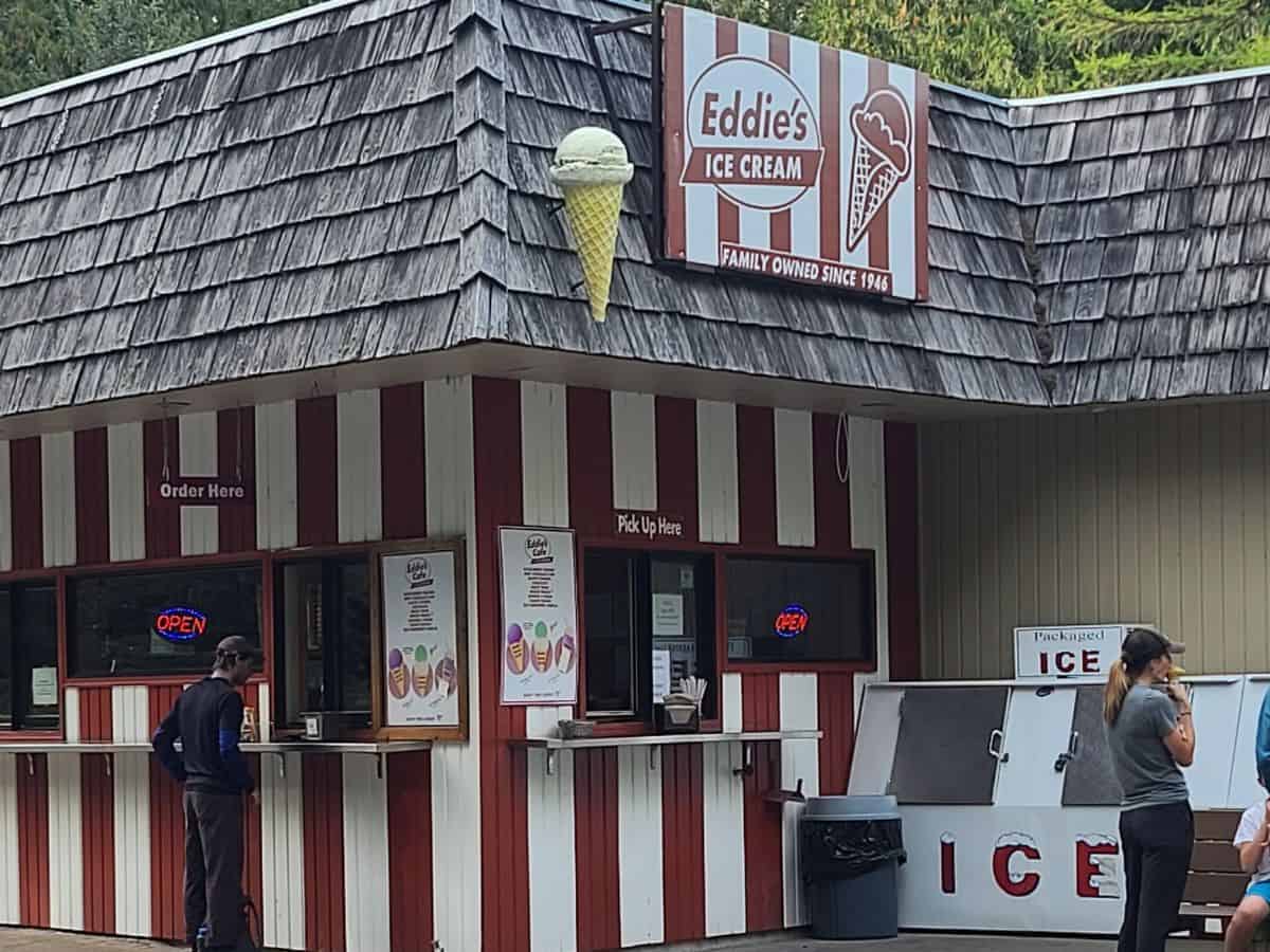 Red and white striped ice cream stand with a sign that reads "Eddie's Ice Cream Family Owned Since 1960" and a large cone decoration on the roof. People stand at the service window and beside a freezer labeled "ICE" at the popular snack stop in Glacier National Park featured in a Glacier National Park itinerary.