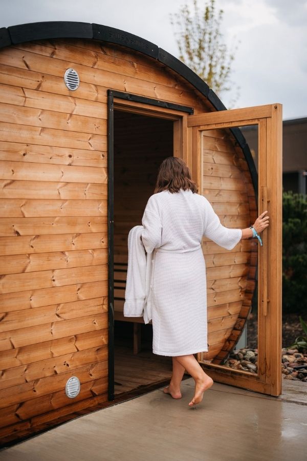 Person in a white robe steps into a wooden barrel sauna, holding a towel beside the open door. This relaxing wellness experience offers a unique way to unwind and recharge during a Zion National Park itinerary.