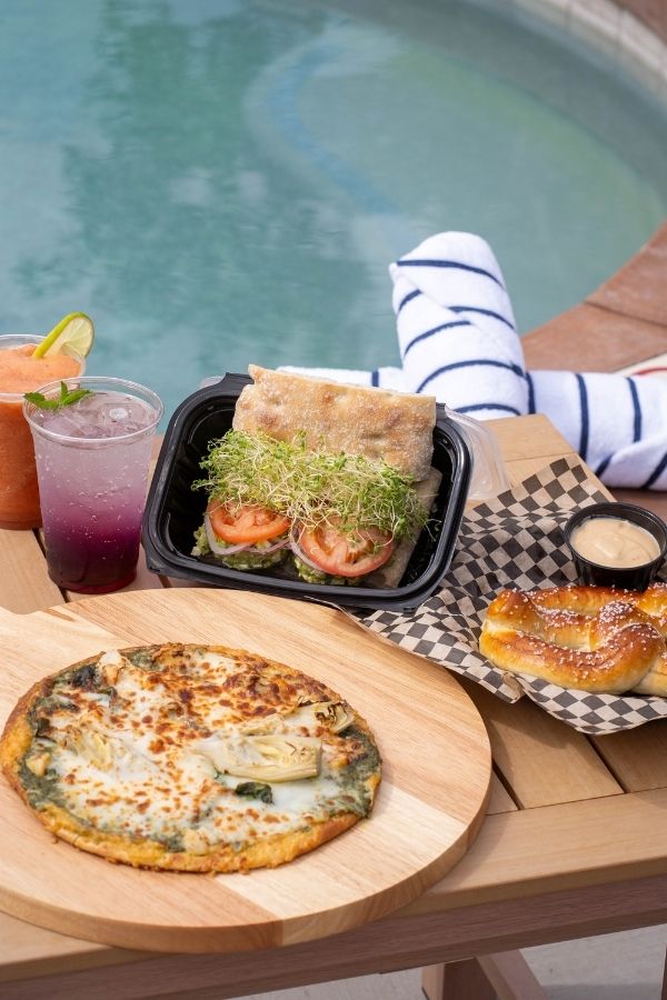 Poolside spread with a flatbread pizza, sandwich topped with sprouts and tomatoes, soft pretzel with dipping sauce, and two colorful drinks on a wooden table beside the water. This casual dining scene highlights a relaxing meal stop to include in a Zion National Park itinerary after a day of exploring.