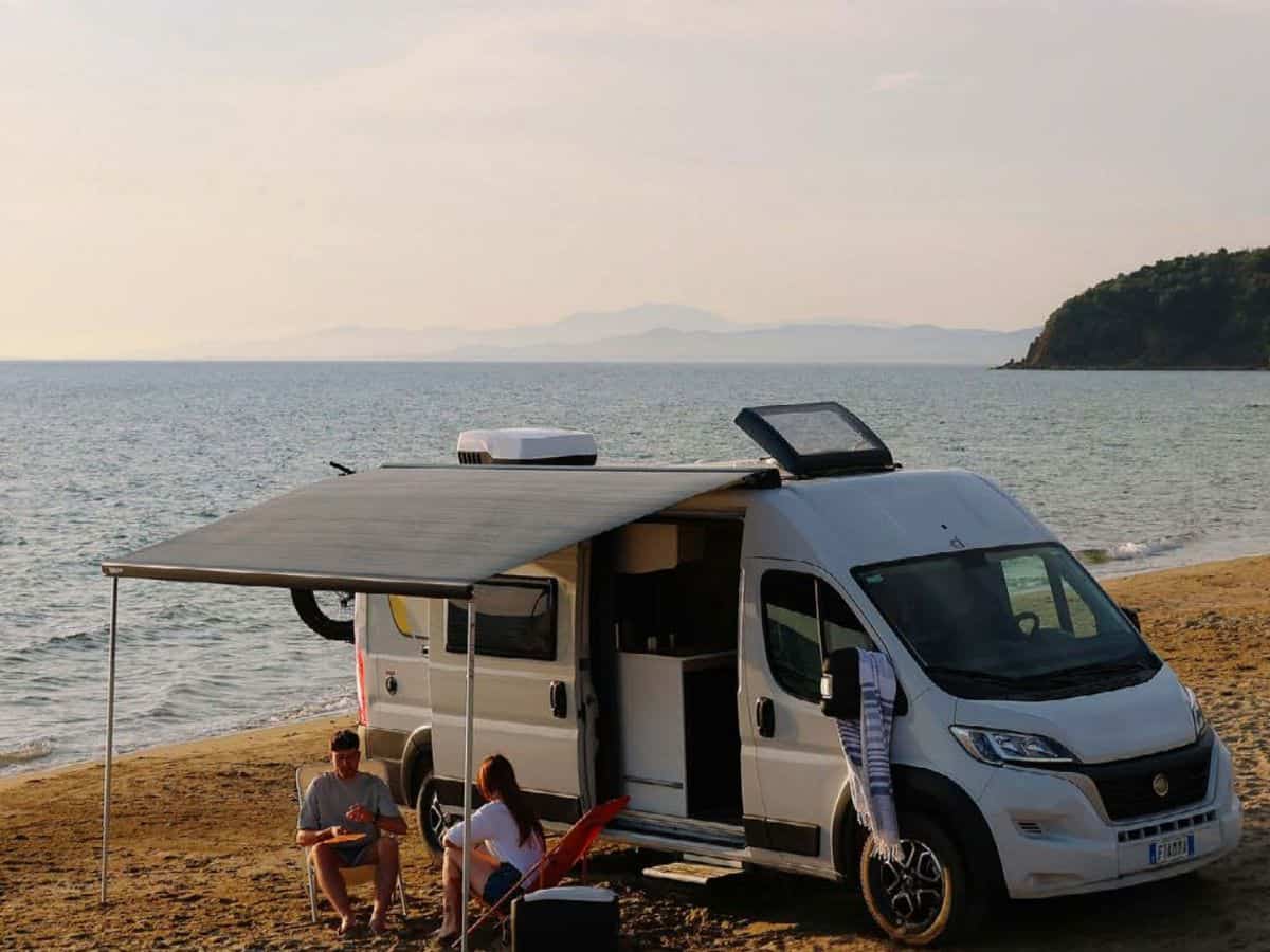Camper van parked on a sandy beach with an extended awning providing shade over two people sitting in folding chairs by the water. Scene shows a relaxed coastal setup demonstrating how a Fiamma F45S or F80S awning creates outdoor living space beside a van.