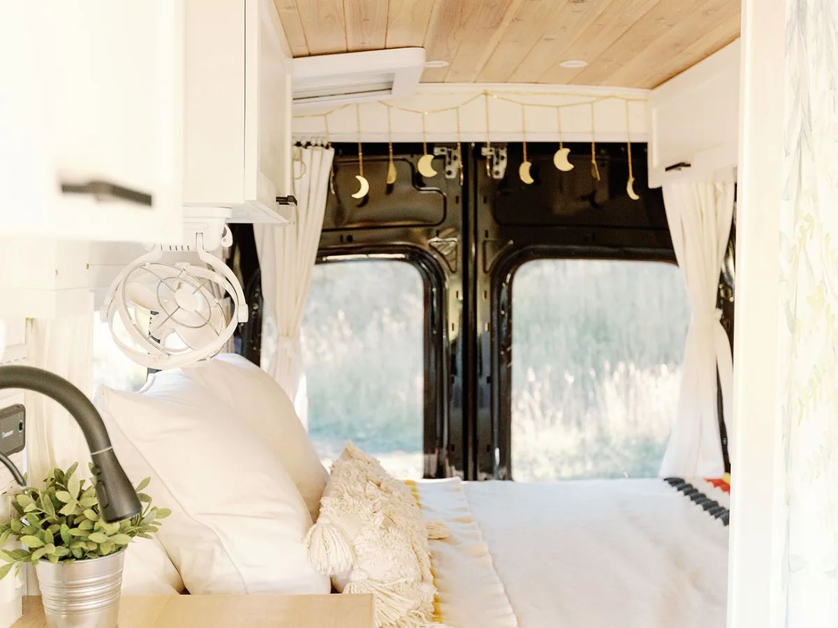 MaxxAir Fan Deluxe Review Bright and airy camper van interior with white bedding and wood-paneled ceiling, featuring a MaxxAir Fan Deluxe installed above the bed to provide ventilation and airflow for van life comfort.