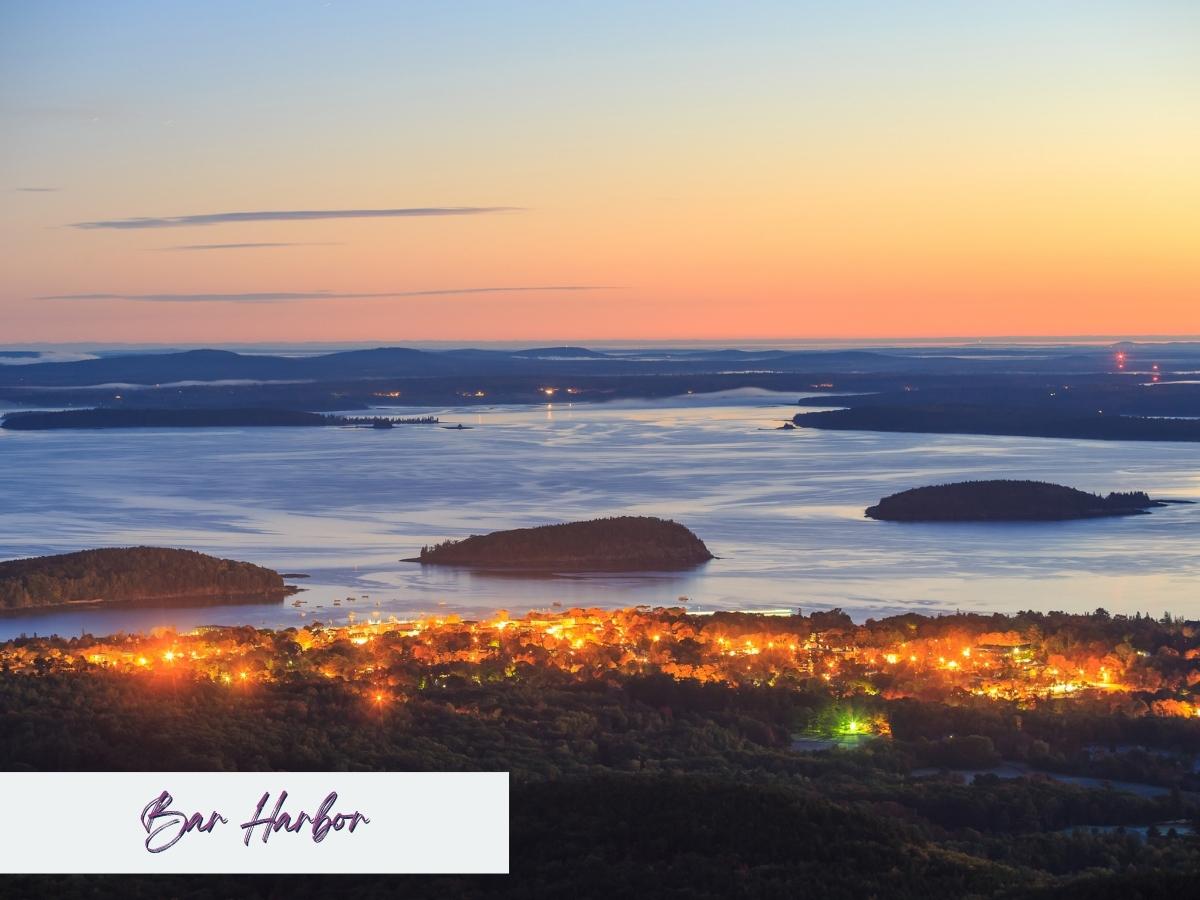 Twilight view of Bar Harbor in the fall near Acadia National Park in October, with the town glowing warmly against the backdrop of calm ocean waters and scattered islands. The sky fades from orange to deep blue as night falls over the autumn landscape.