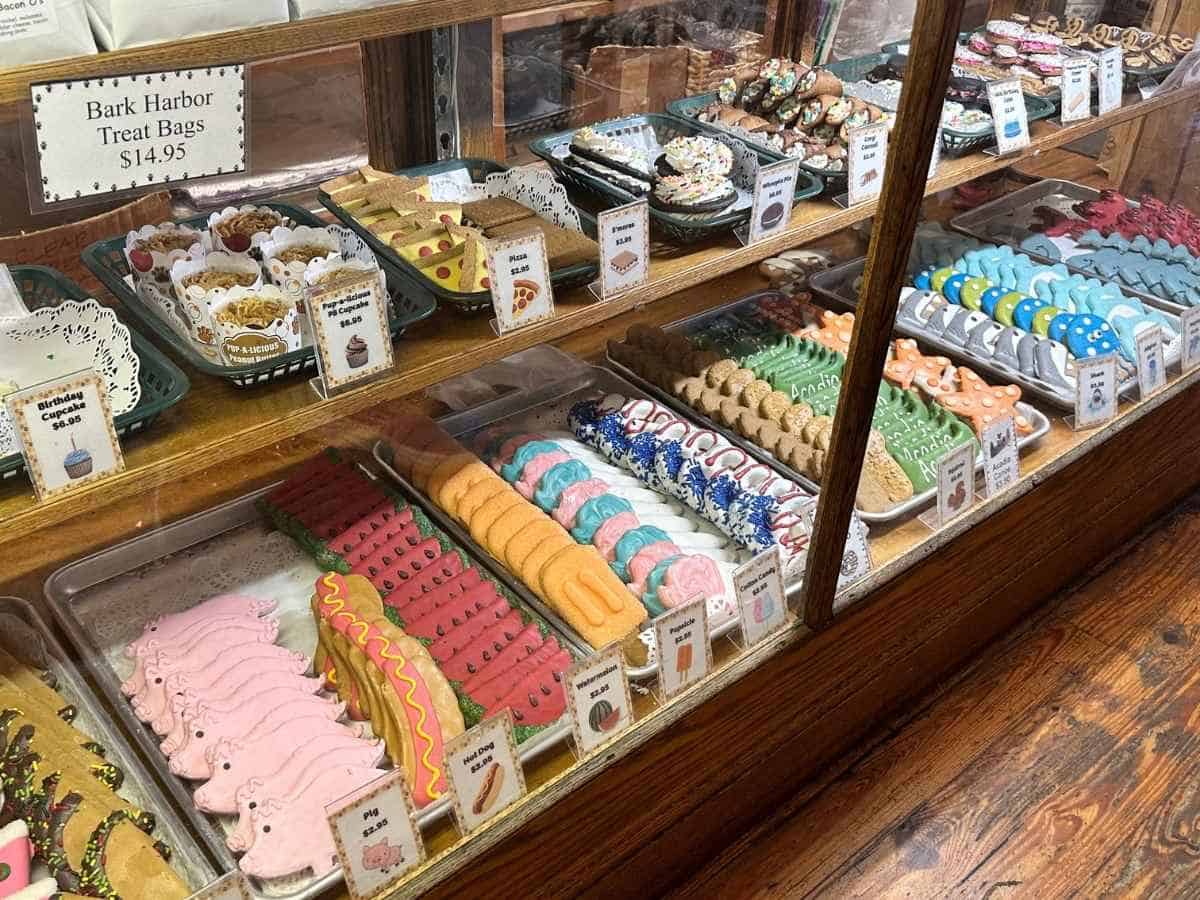 are dogs allowed in Acadia National Park? A bakery display case is filled with colorful dog treats shaped like pigs, hot dogs, bones, and other fun designs, with a sign reading “Bark Harbor Treat Bags \$14.95.” This image complements “Are dogs allowed in Acadia National Park” by highlighting Bar Harbor Maine pet-friendly shops catering to visiting dogs.