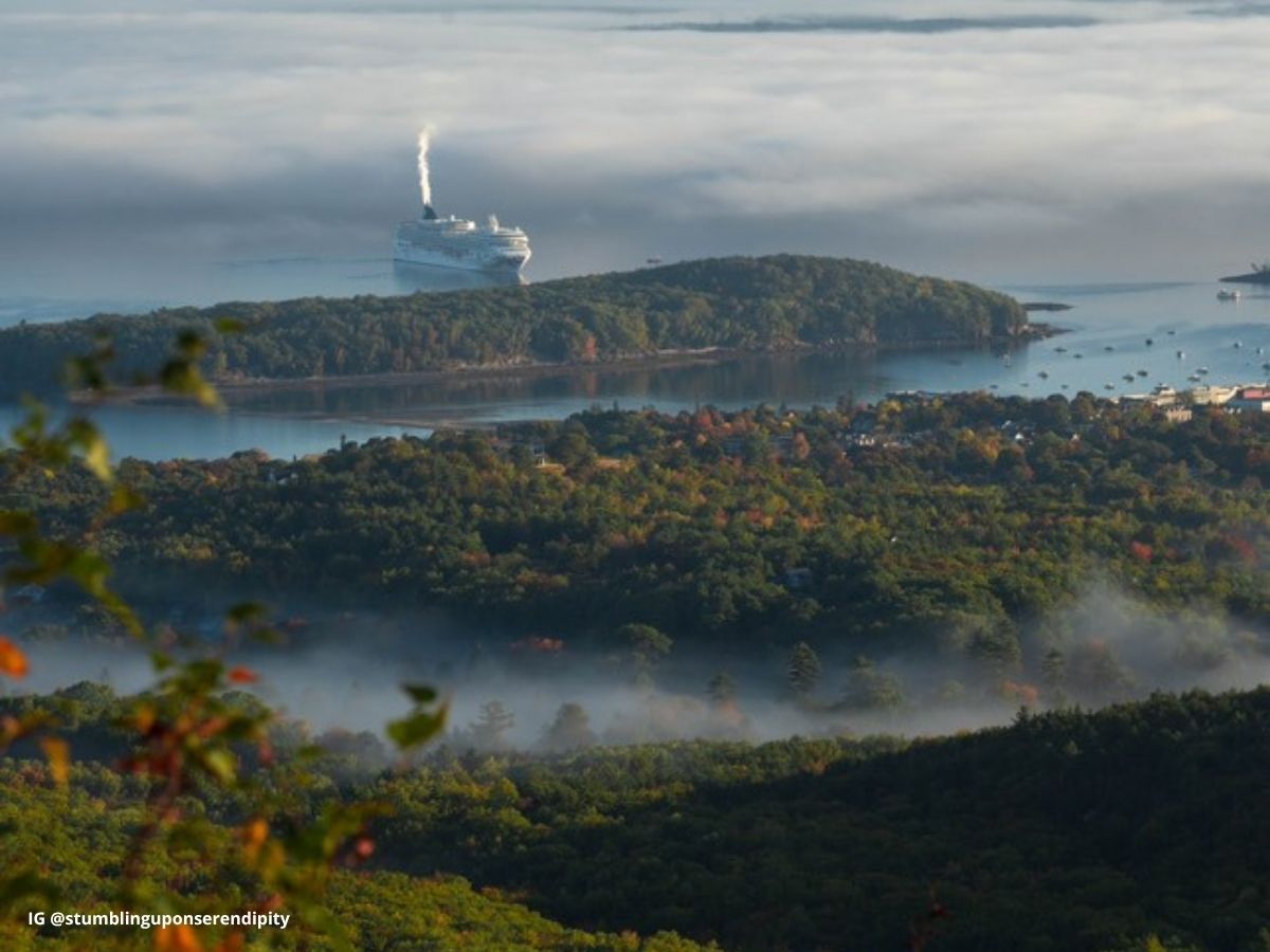 Cruise ship approaching Bar Harbor near Acadia National Park in the fall, with mist rising over forested hills and colorful autumn foliage. Small boats and coastal town structures are visible in the harbor under a foggy morning sky.