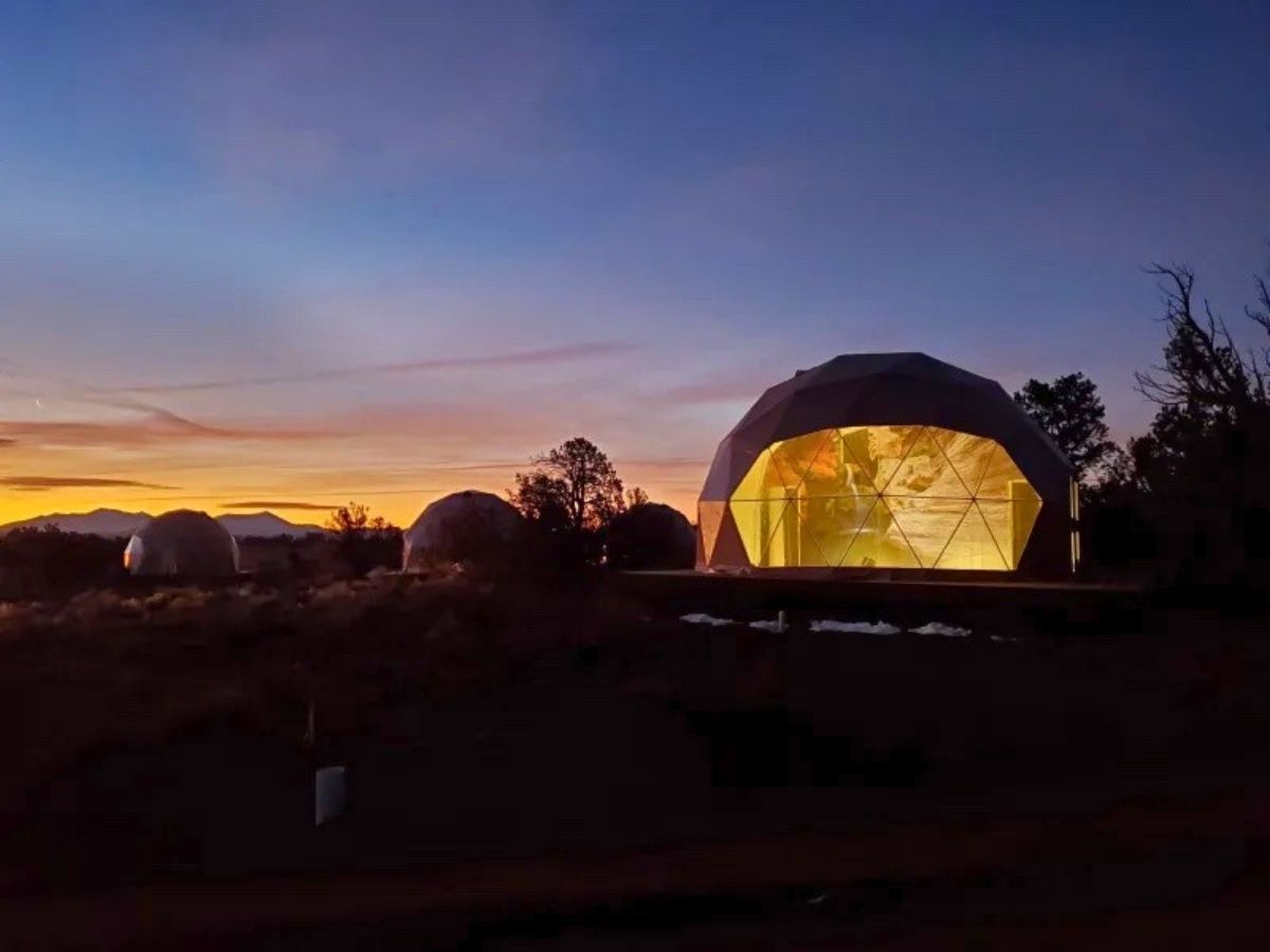 Geodesic dome glamping tents glow warmly at sunset in a desert landscape near Bryce Canyon with silhouettes of trees and distant mountains on the horizon. The sky fades from orange to deep blue creating a peaceful evening atmosphere around the domes. This unique stay option highlights glamping as part of a Bryce Canyon itinerary.
