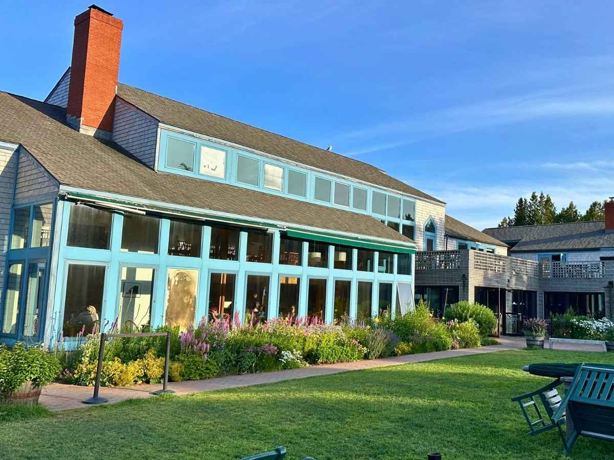 Jordan Pond House coastal lodge in Acadia National Park in the fall features light blue-trimmed windows, blooming flower beds, and a spacious lawn with Adirondack chairs. The warm evening sunlight highlights the building’s shingle siding and brick chimneys against a clear sky.