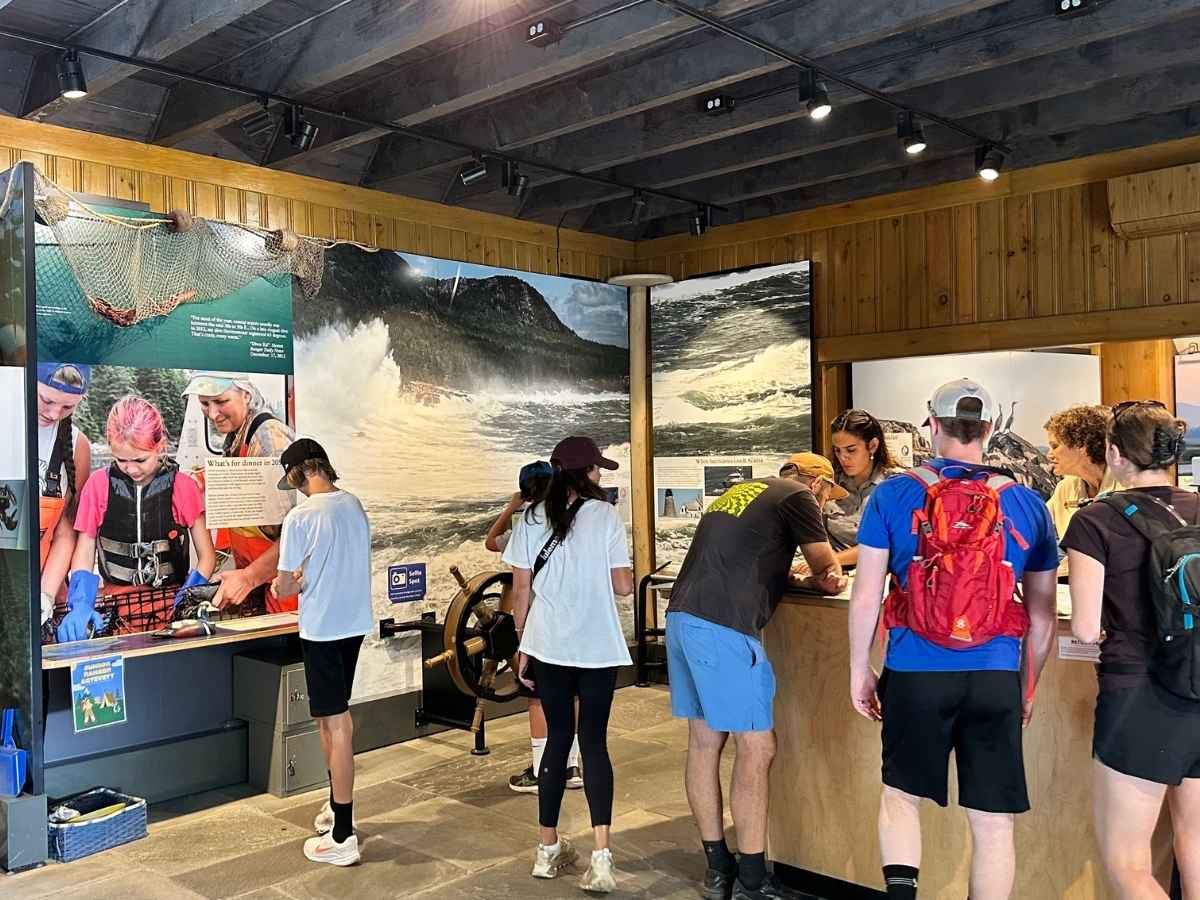 Visitors gather inside a nature center at Acadia National Park in the fall, exploring educational exhibits about marine life and coastal ecosystems. A ranger assists guests at the desk while colorful wall displays and hands-on activities engage families and hikers.
