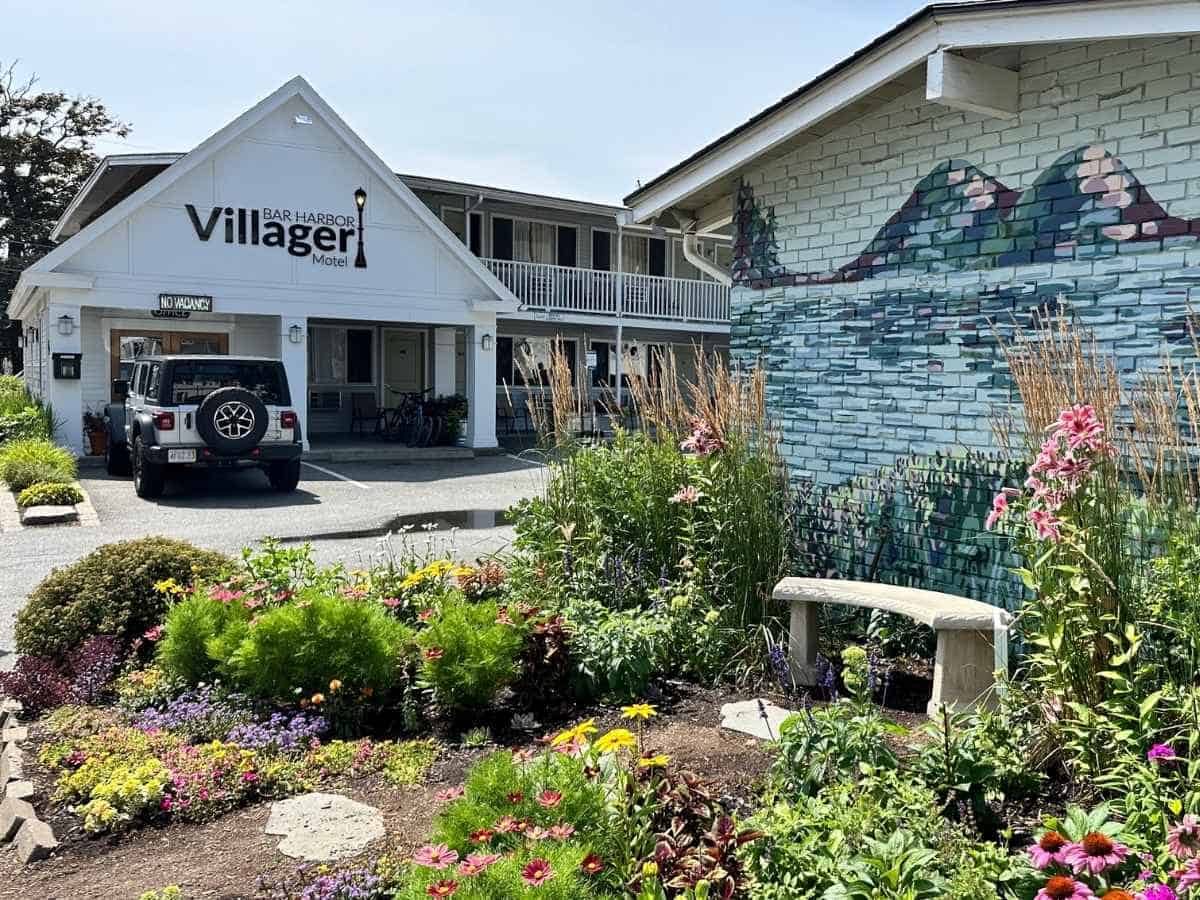 The Bar Harbor Villager Motel displays a crisp white facade with a “No Vacancy” sign and a parked Jeep out front, while a colorful mural of Acadia's mountain landscape is painted on an adjacent brick wall. In the foreground, vibrant wildflowers and a small stone bench create a peaceful garden scene, capturing the charm of lodging near Acadia National Park in the fall.