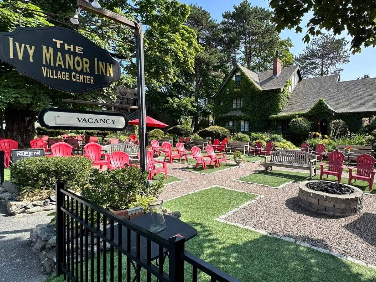 Ivy Manor Inn in Bar Harbor Maine, features a lush ivy-covered building surrounded by tall trees and a neatly manicured courtyard filled with red Adirondack chairs and a central stone fire pit. The setting is welcoming and peaceful, making it an inviting lodging option near Acadia National Park in the fall.