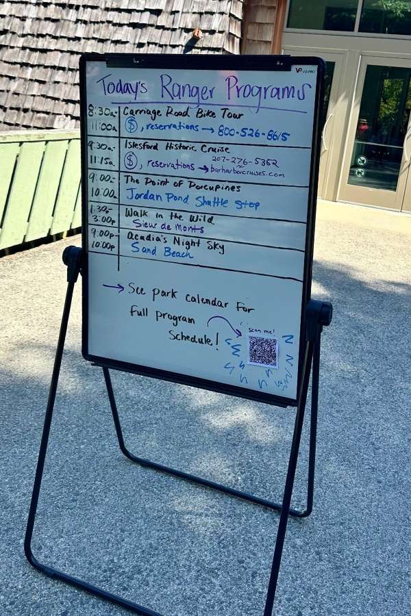 A whiteboard outside the Hulls Cove visitor center at Acadia lists the day’s ranger programs at Acadia National Park in the fall, including events like the Carriage Road Bike Tour, Islesford Historic Cruise, and Acadia’s Night Sky. A QR code directs visitors to the full schedule, with handwritten notes and times adding a personal touch.