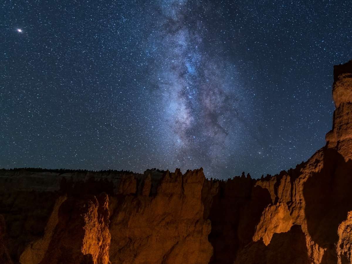 Milky Way stretches across a star filled night sky above the jagged hoodoos of Bryce Canyon, illuminated by warm light against the dark landscape. Tall rock formations rise from the canyon floor creating dramatic silhouettes beneath the galaxy. This nighttime view highlights stargazing as a memorable experience on a Bryce Canyon itinerary.