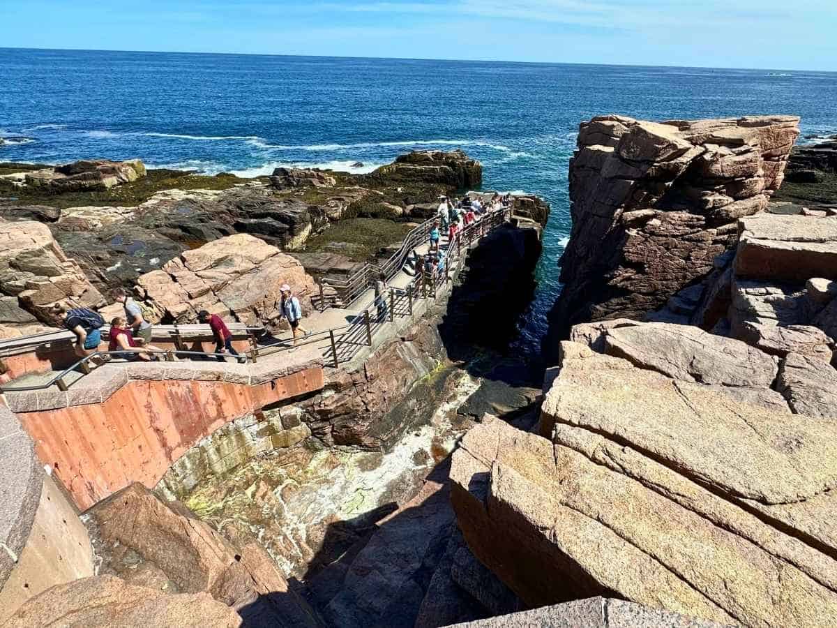 Tourists walk along a rocky cliffside path overlooking the dramatic coastal feature known as Thunder Hole in Acadia National Park in the fall. Surrounded by massive pink granite formations and crashing waves, the scene captures the park’s rugged Atlantic shoreline under a bright blue sky.