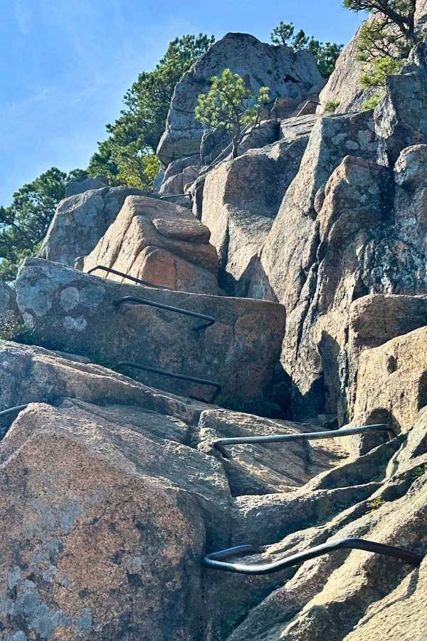 A steep granite cliff on Beehive hike in Acadia National Park in the fall is fitted with a series of black iron rungs, forming a challenging climbing route. Jagged rock faces and scattered pine trees rise toward a bright blue sky.