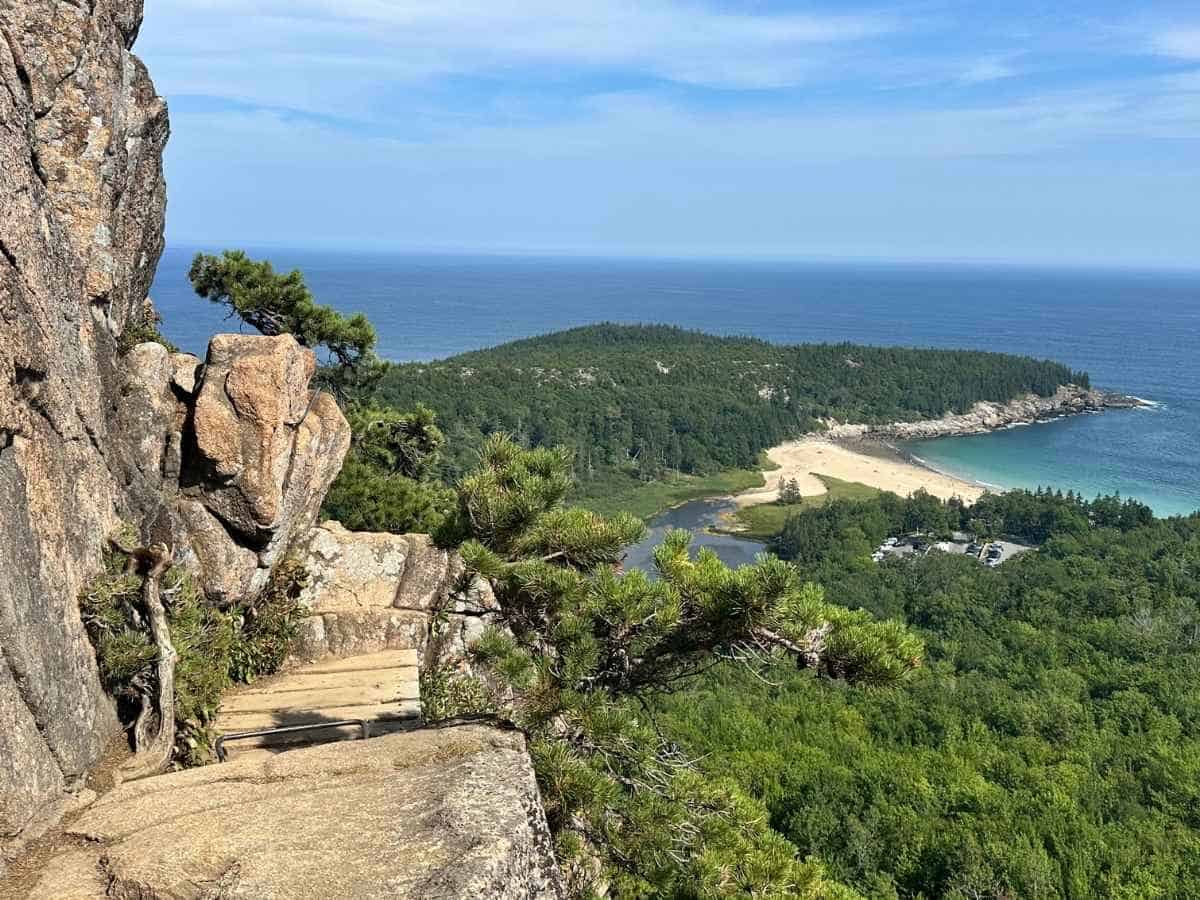 A dramatic cliffside view in Acadia National Park in the fall overlooks Sand Beach, where turquoise waves meet golden sand surrounded by dense evergreen forest. Stone steps built into the rocky trail add to the rugged beauty of the coastal landscape.