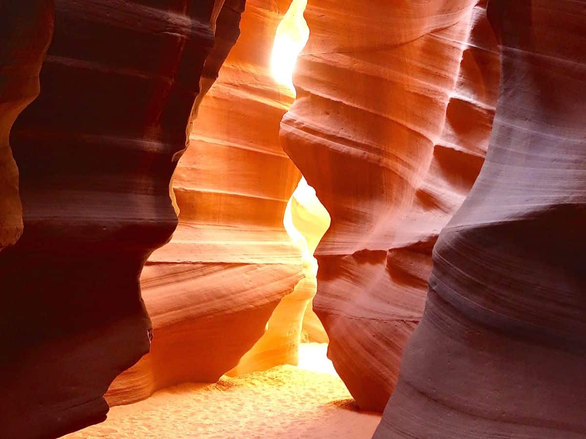 Narrow sandstone walls of a glowing slot canyon with warm orange and red tones lit by sunlight streaming through the opening above. Curved rock formations create smooth flowing shapes along a sandy pathway, showcasing a scenic stop often included on a route from Page to Grand Canyon National Park. Light and shadow highlight the depth and texture of this iconic desert landscape.
