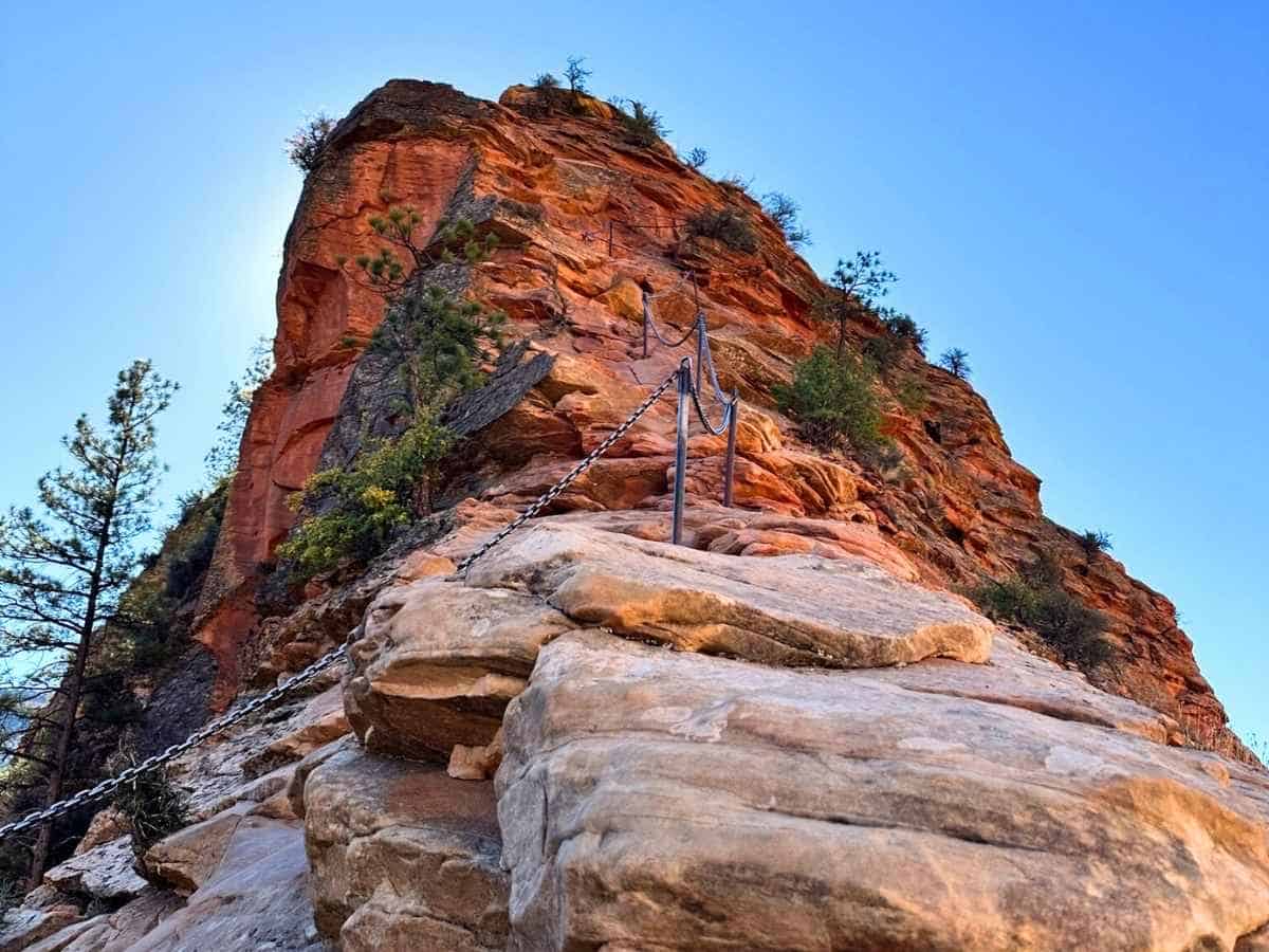 Steep red rock peak with a narrow trail secured by metal chains, a thrilling viewpoint along a las vegas to zion national park road trip. The rugged sandstone formation rises against a clear blue sky, highlighting one of Zion’s most iconic and adventurous hikes.