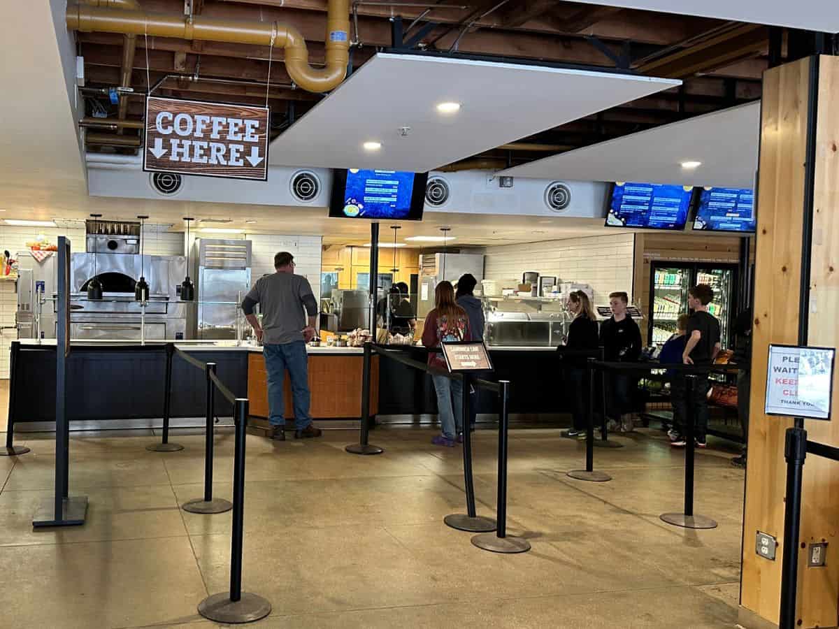 Interior of Degnan’s Kitchen in Yosemite Valley, showing visitors ordering food at a modern counter beneath a rustic "Coffee Here" sign, with menu screens and a pizza oven in the background. A convenient breakfast or lunch stop during a one day in Yosemite National Park itinerary.