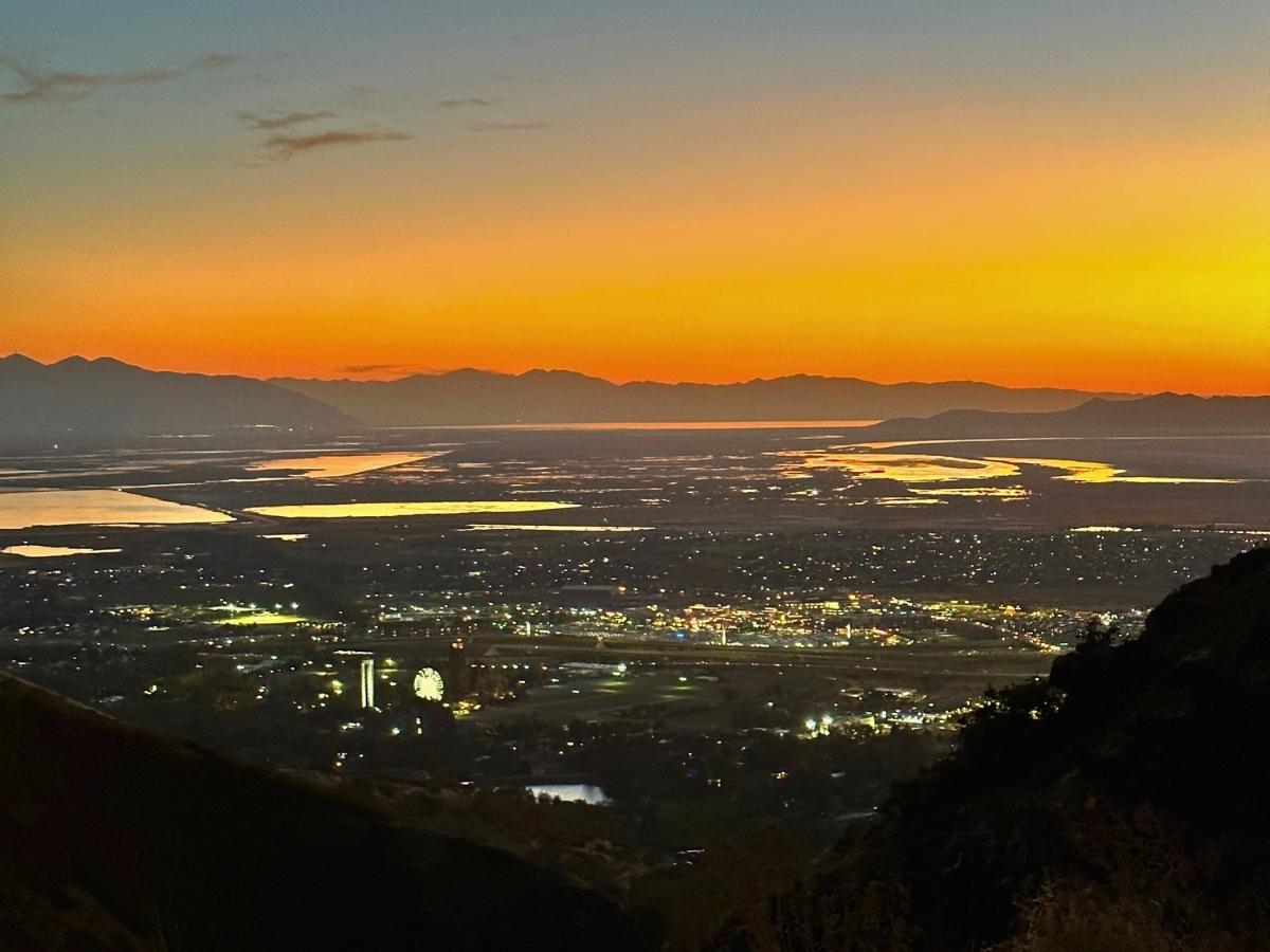 City skyline at dusk with glowing lights below and reflective water winding through the landscape under a vibrant orange sunset sky. Scenic starting point for the drive from Salt Lake City to Arches National Park road trip, capturing the city and surrounding mountains at golden hour.