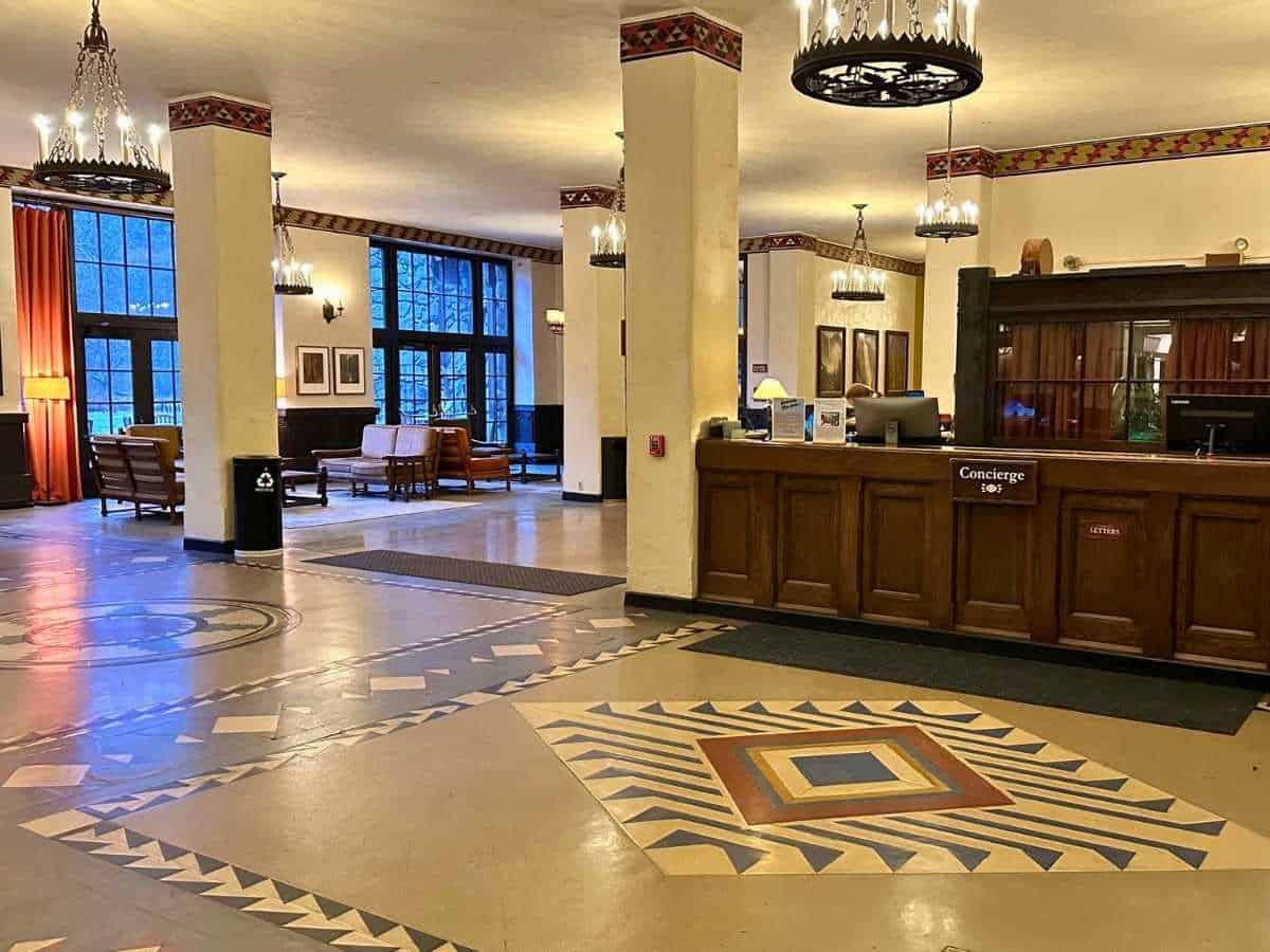 The historic Ahwahnee hotel lobby features a wooden concierge desk, patterned tile floors, and tall columns with decorative trim. Large windows, chandeliers, and cozy seating areas create a warm and inviting stay to consider in a Yosemite itinerary.