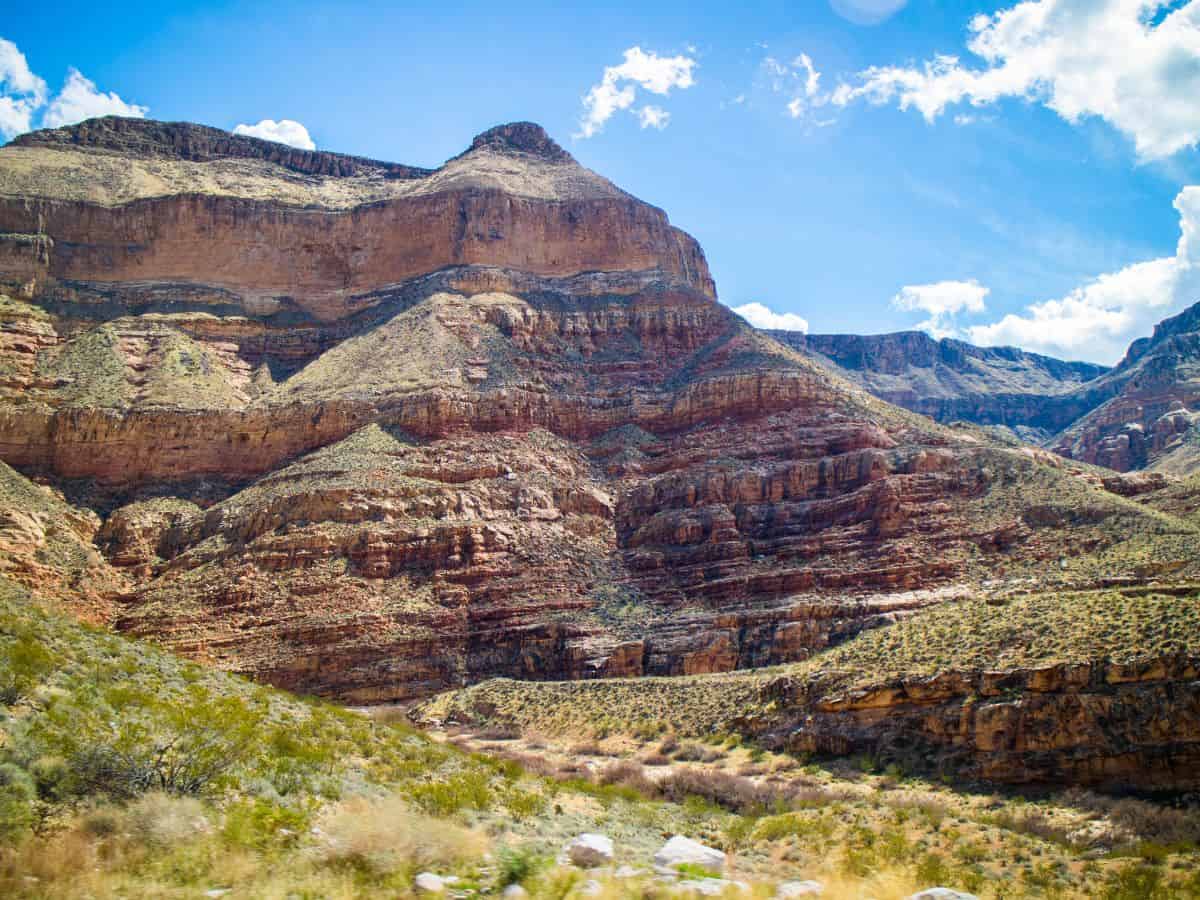 Layered desert cliffs and mesas rising beneath a bright blue sky, showcasing the rugged terrain along a las vegas to zion national park road trip. The striped rock formations and scattered desert vegetation highlight the dramatic Southwest landscape travelers experience on the drive to Zion.