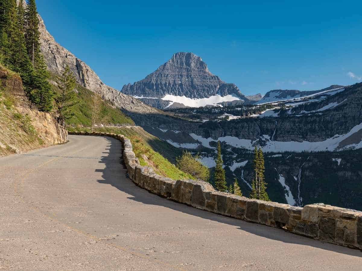 Curving section of Going-to-the-Sun Road with a stone guard wall leading toward a sharp mountain peak with patches of snow in Glacier National Park. The scenic alpine drive highlights one of the most iconic viewpoints included in a Glacier National Park itinerary.