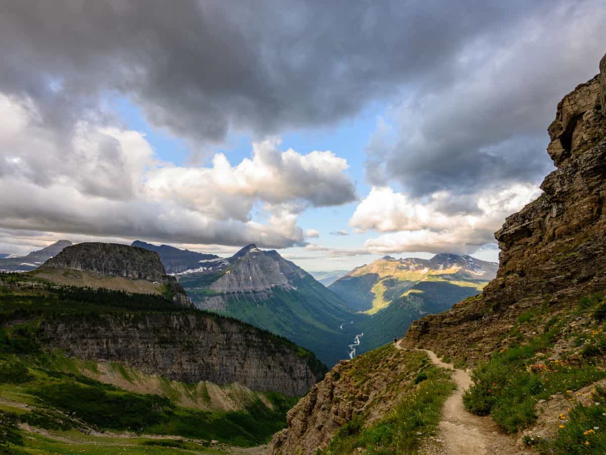 Narrow dirt path along a cliff’s edge on the Highline Trail in Glacier National Park, with dramatic mountain views, a winding valley river, and moody clouds—highlighting one of the best hikes in Glacier National Park.