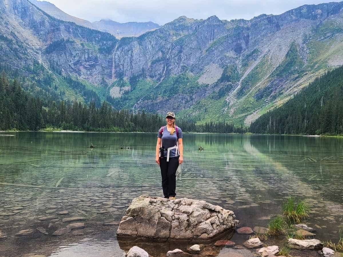 Woman standing on a rock at the edge of Avalanche Lake with clear water revealing stones and submerged logs beneath the surface. Steep mountains and evergreen forest rise behind the lake with thin waterfalls visible on the cliffs in Glacier National Park as featured in a Glacier National Park itinerary.