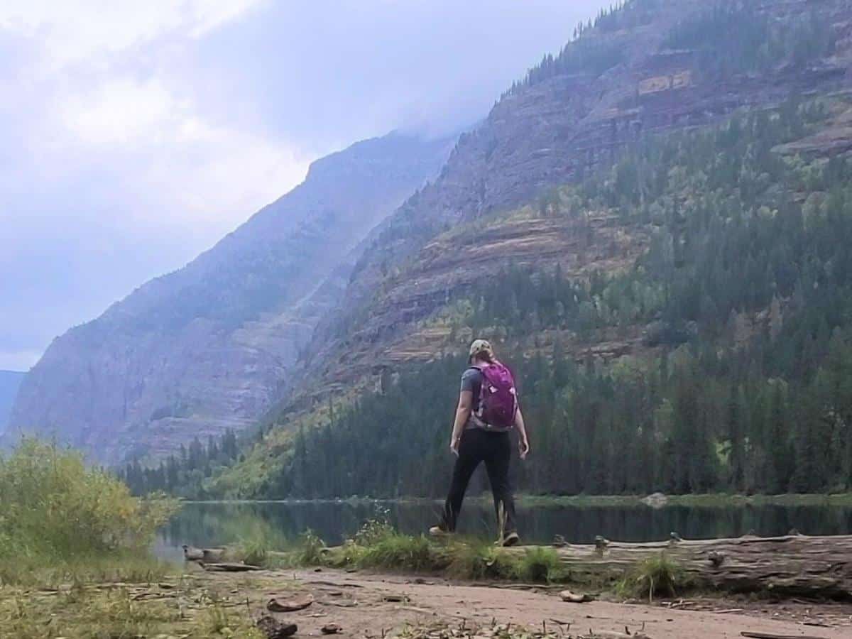 Hiker with a purple backpack walking along the rocky shore of Lake McDonald with calm water reflecting forested mountains in Glacier National Park. Layered cliffs and evergreen trees rise above the lake under cloudy skies as featured in a Glacier National Park itinerary.