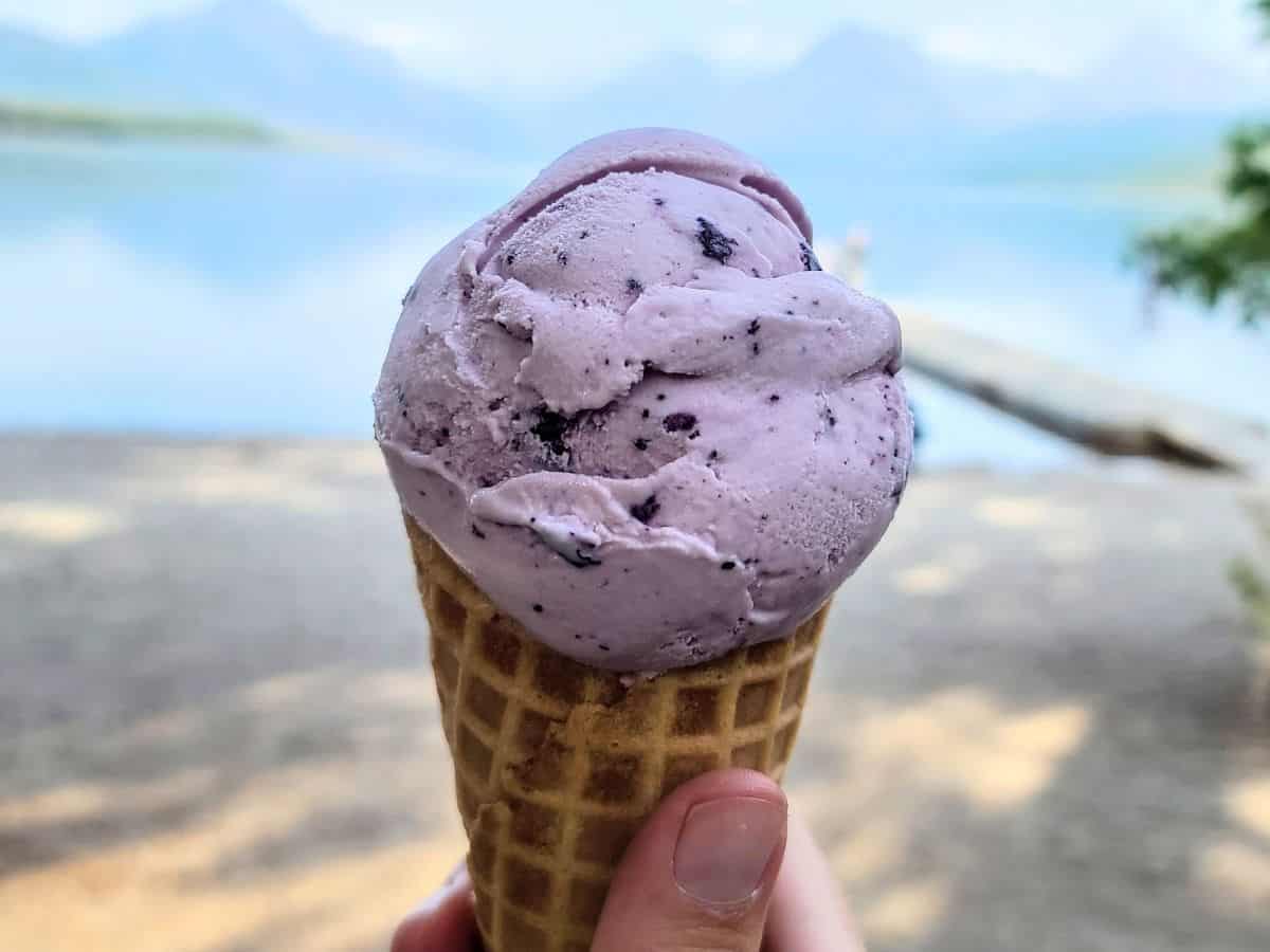 Hand holding a waffle cone with a scoop of purple berry ice cream in front of the shoreline of Lake McDonald in Glacier National Park. Calm blue water and hazy mountains appear in the background as a treat stop during a Glacier National Park itinerary.