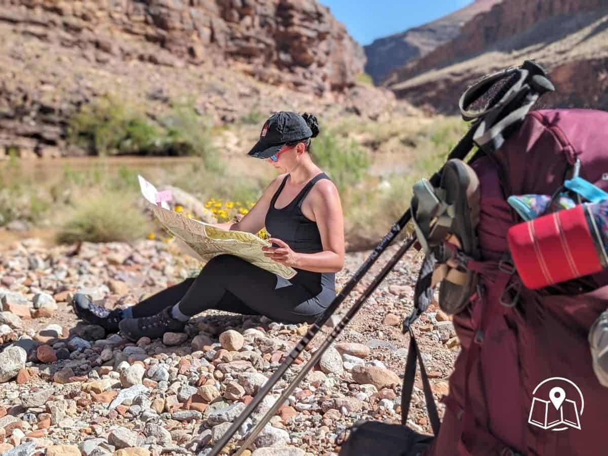 A woman dressed in black athletic gear sits on a rocky trail in the Grand Canyon, studying a map under the bright sun with trekking poles and a large backpack nearby. This candid moment captures a pause in an adventurous Grand Canyon itinerary, emphasizing self-guided exploration and rugged terrain.