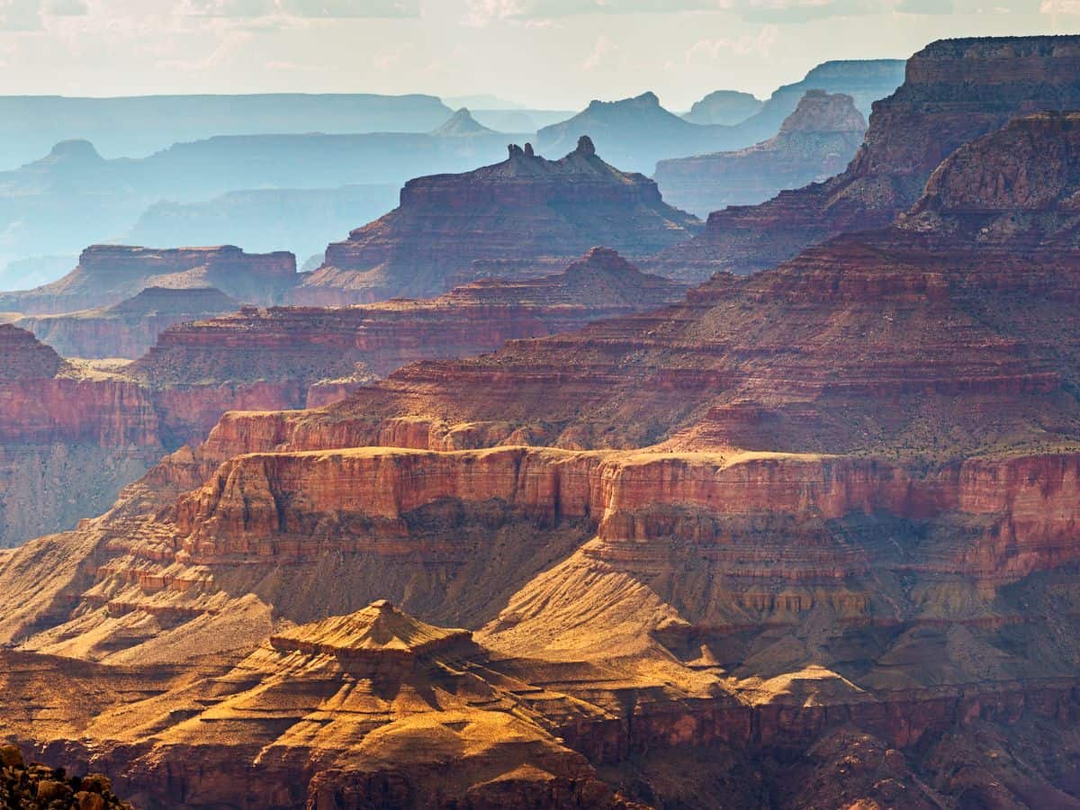 Layered rock formations of the Grand Canyon bathed in warm sunlight with hazy blue ridges fading into the distance. A breathtaking finale to a road trip from Las Vegas to Grand Canyon by car.