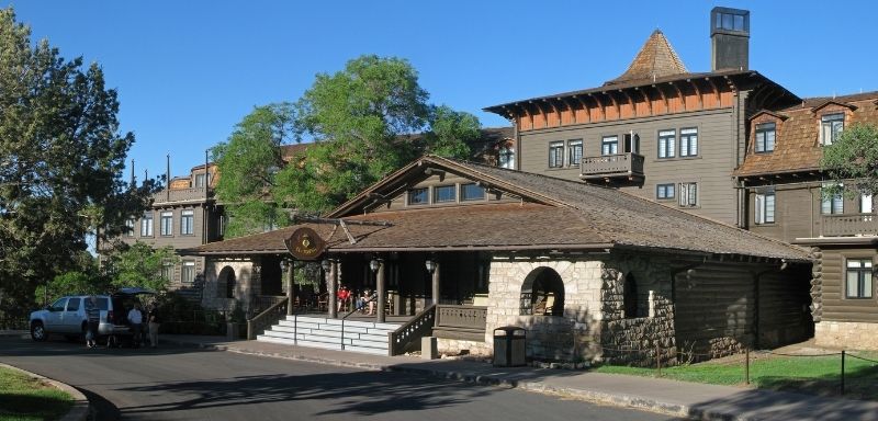 Color photo of the El Tovar Hotel at the South Rim of the Grand Canyon, featuring rustic stone and wood architecture with guests gathered near the shaded front porch. This historic lodge is a popular overnight stop for travelers planning a classic Grand Canyon itinerary.