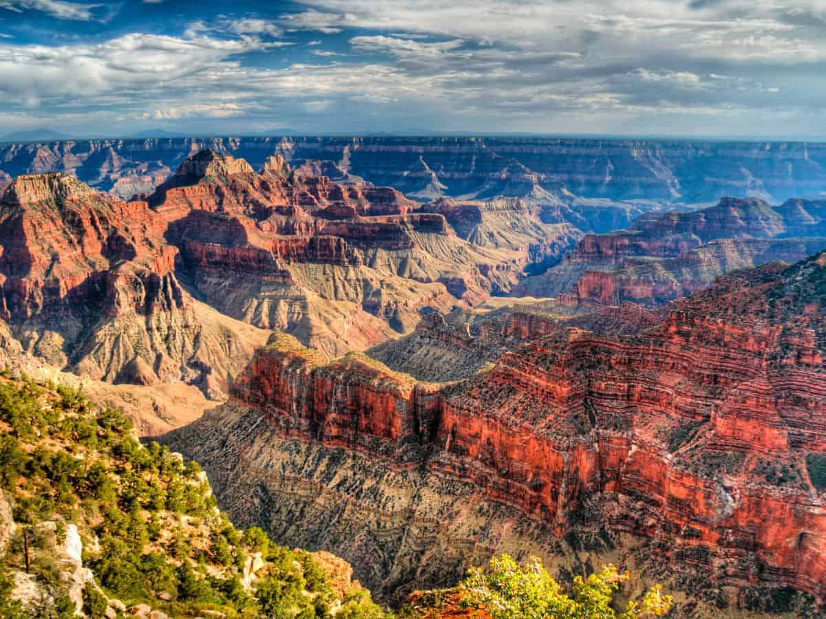 Vibrant, high-contrast view of the Grand Canyon’s layered red rock formations and sweeping ridgelines under a partly cloudy sky. This iconic landscape showcases the dramatic scenery that draws travelers to build a Grand Canyon itinerary around hiking, sightseeing, and photography.