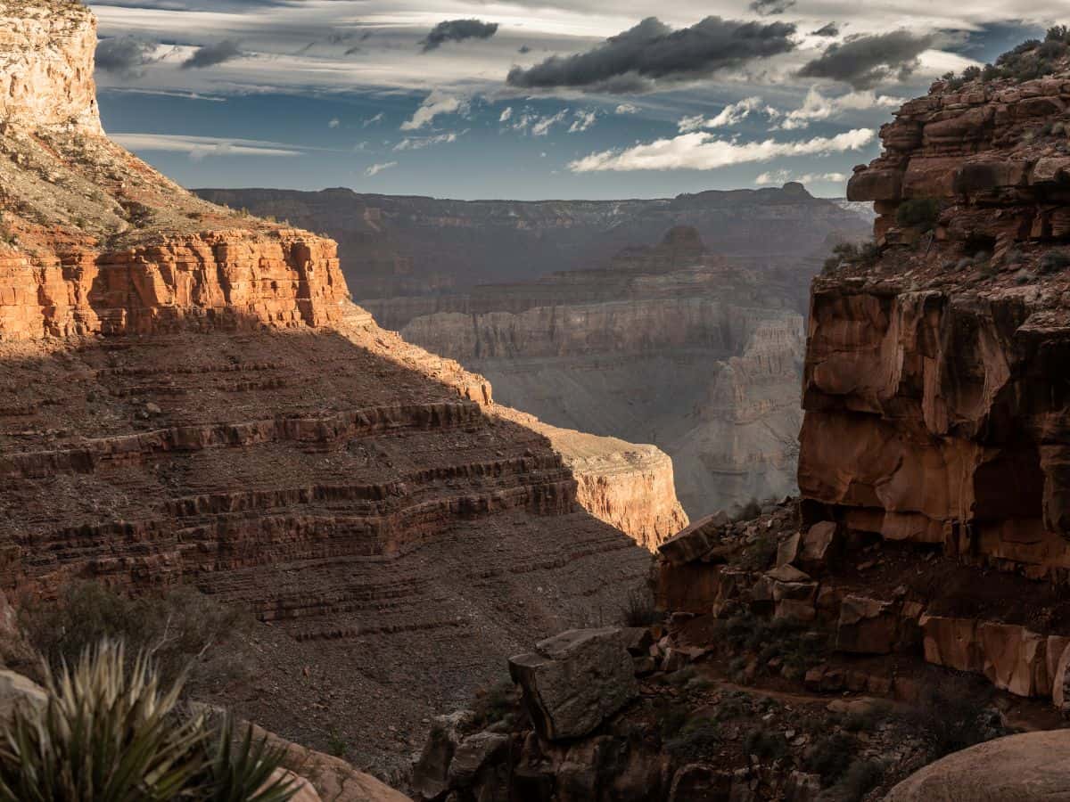 Moody view of the Grand Canyon’s steep, layered cliffs under a dramatic sky, with pockets of golden light hitting the rock formations while deep shadows fill the valleys. This atmospheric scene captures the awe-inspiring scale and shifting beauty that make the canyon a key destination on any Grand Canyon itinerary.