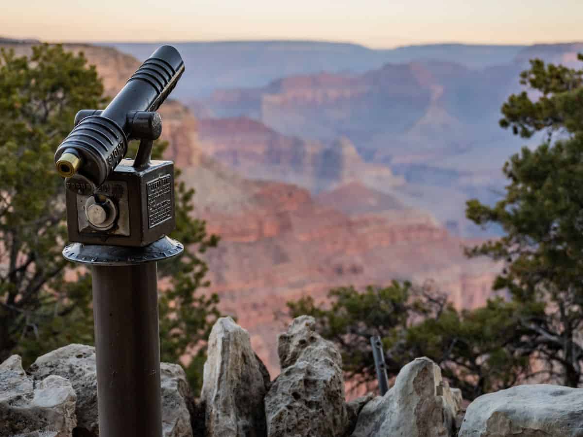 A coin-operated scenic viewer is mounted at Hermits Rest overlook, pointed toward a hazy expanse of colorful canyon walls and ridges at dusk. This viewpoint invites travelers on a Grand Canyon itinerary to pause and take in the vast landscape in greater detail.