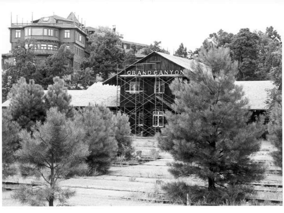 Black and white photo of the historic Grand Canyon train depot surrounded by pine trees, with the words "Grand Canyon" visible above the central entrance under scaffolding. In the background, the iconic El Tovar Hotel sits perched on a hilltop, adding historic charm and relevance to any Grand Canyon itinerary.