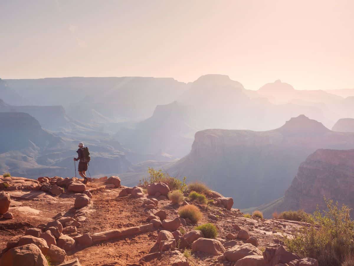 A lone hiker with trekking poles walks along a red dirt trail at sunrise or sunset, surrounded by the hazy, layered silhouettes of the Grand Canyon’s vast landscape. This dreamy moment captures the solitude and scale that make hiking a memorable part of any Grand Canyon itinerary.