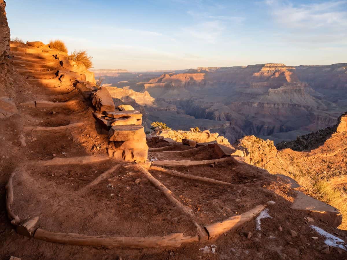 Stone steps carved into the canyon wall glow in warm morning light, leading South Kaibab hikers down a switchback trail with sweeping views of the Grand Canyon’s vast, layered landscape. This rugged descent is a scenic highlight often included in an active Grand Canyon itinerary.