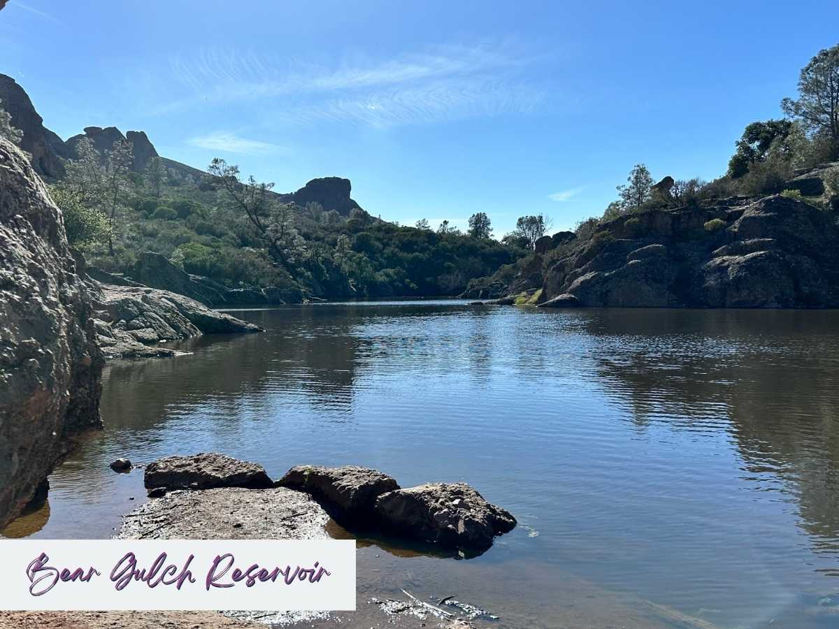 Hiking Pinnacles National Park Itinerary Tranquil view of Bear Gulch Reservoir surrounded by rocky cliffs and lush greenery under a clear blue sky, highlighting the peaceful scenery found on the best hikes in Pinnacles National Park. Text overlay reads "Bear Gulch Reservoir."
