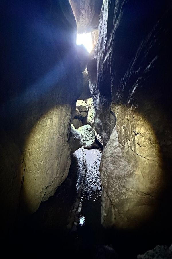 Hiking Pinnacles National Park Itinerary Narrow, dark passage inside Bear Gulch Cave with rocky walls, a small stream, and a shaft of sunlight illuminating the exit, showcasing the unique cave exploration experience hiking Pinnacles National Park.