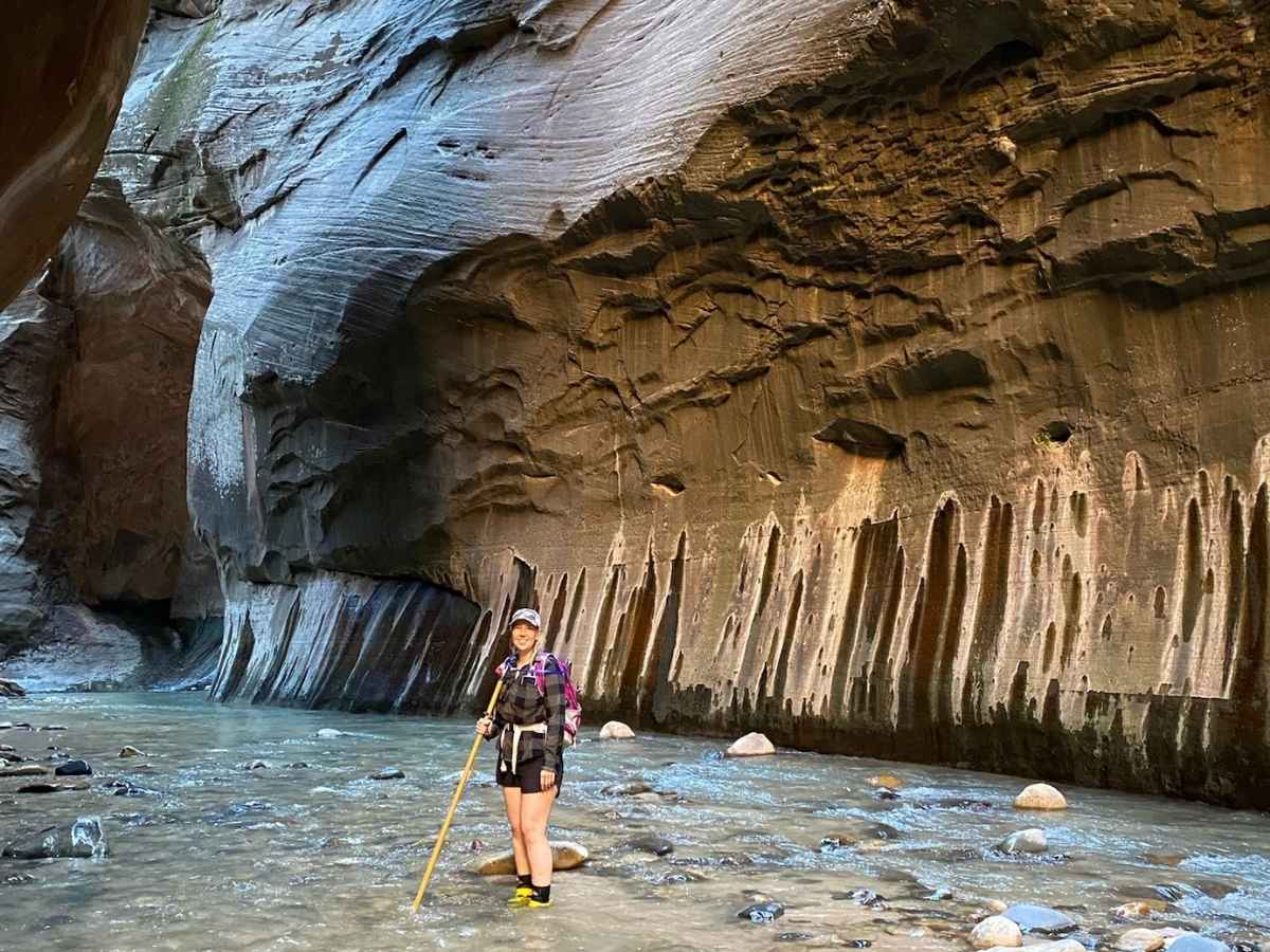 A smiling hiker stands in shallow river water with a walking stick, wearing water shoes, a daypack, and layered hiking gear suitable for The Narrows in Zion National Park. Behind her, steep canyon walls with dramatic vertical streaks emphasize the rugged and wet conditions that make choosing the best gear for hiking The Narrows essential.