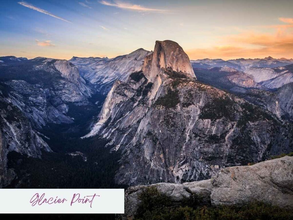 From Glacier Point, Half Dome rises sharply against a pastel Yosemite sunset sky, overlooking the vast, forested valley below. The scene captures dramatic granite formations and the layered beauty of the Sierra Nevada as day fades to dusk.