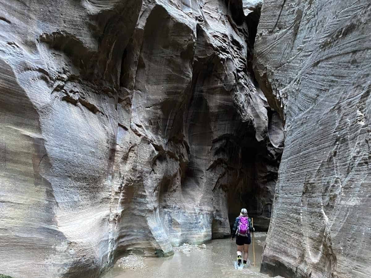 A hiker with a bright pink backpack and walking stick wades through the narrow slot canyon of The Narrows in Zion National Park, surrounded by towering, striated sandstone walls. The scene captures the dramatic rock formations and shallow Virgin River path typical of hiking The Narrows in Zion National Park.