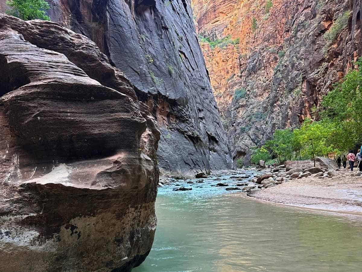 Large, layered boulder known as House Rock sits beside the Virgin River in The Narrows at Zion National Park, with towering canyon walls and hikers visible on the sandy trail to the right. The scene marks a key landmark for those hiking The Narrows, showcasing the blend of natural sculpture and rugged terrain.