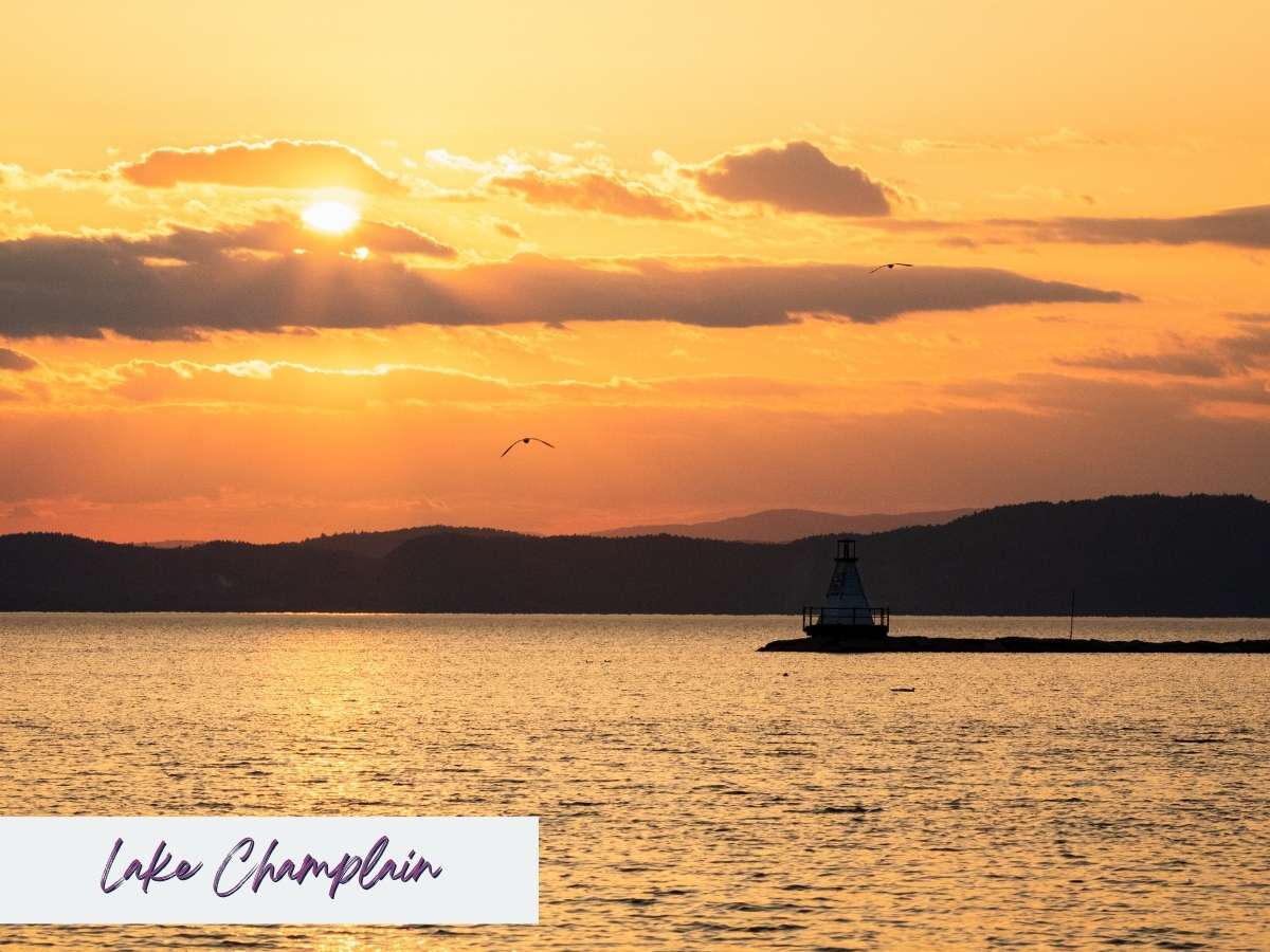 Golden sunset over Lake Champlain with silhouetted mountains and a small lighthouse on the water, offering a peaceful evening view during a 7 day New England road trip itinerary.