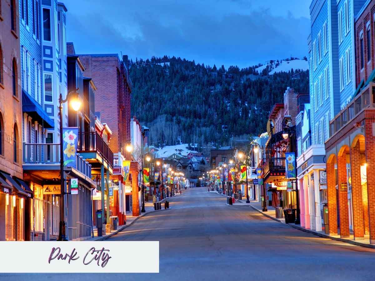 Colorful buildings line historic Main Street in Park City at dusk, with warm lights glowing against a backdrop of snow-dusted mountains — a favorite year-round destination for a cozy day trip from Salt Lake City.