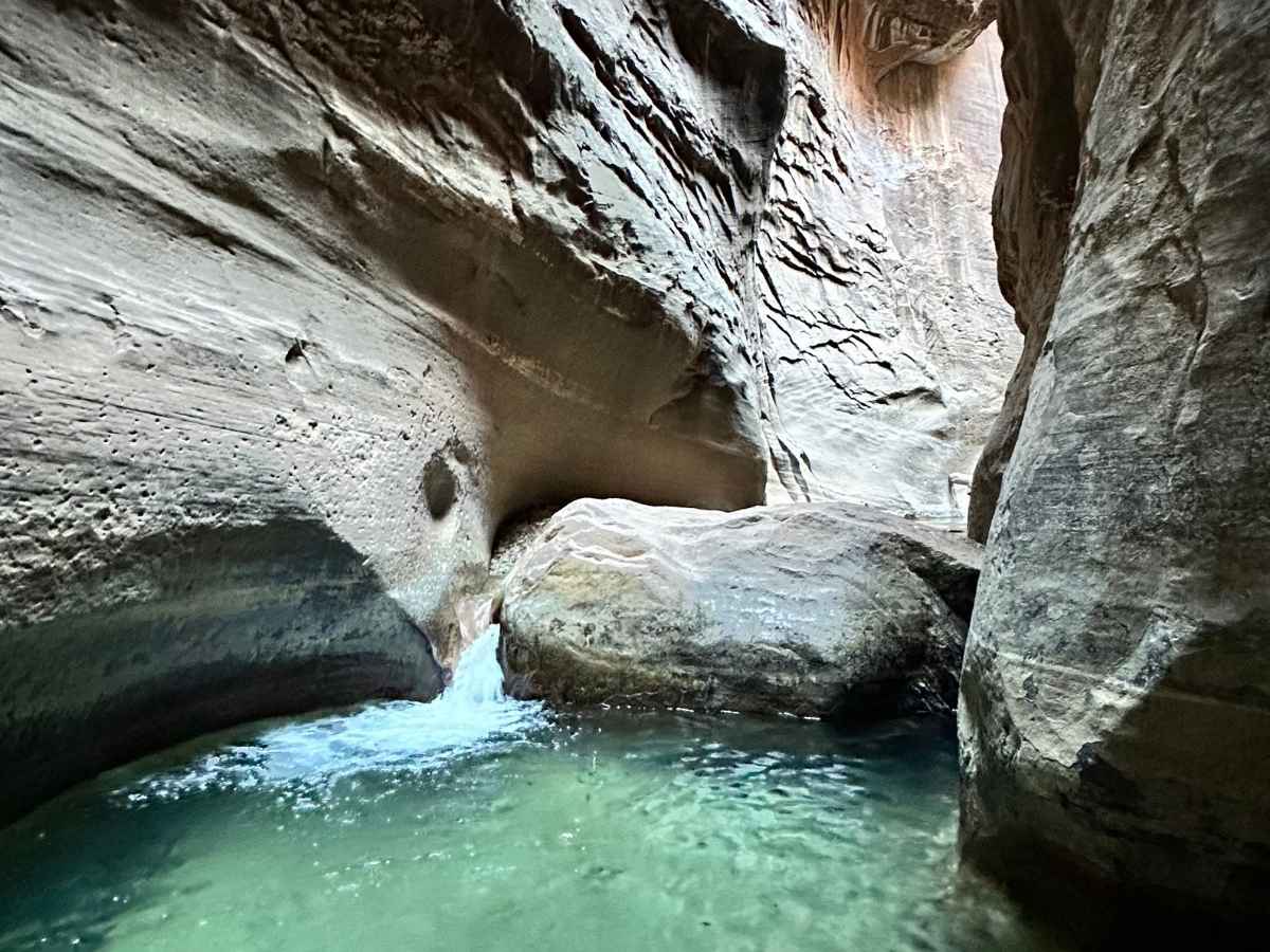 Crystal-clear turquoise water pools at the base of Veiled Falls, where a gentle cascade flows between smooth sandstone walls in The Narrows of Zion National Park. This hidden gem offers a tranquil moment along the hike, with soft lighting and dramatic rock textures creating a secluded canyon oasis.