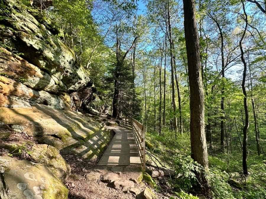 A wooden boardwalk winds along a moss-covered rock ledge through a sunlit forest in Cuyahoga Valley National Park. Hiking this scenic trail is a must on any best things to do in Cuyahoga Valley National Park itinerary.