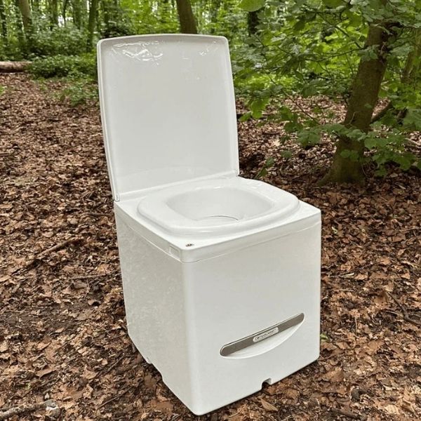 White Compo Closet Cuddy composting toilet with lid open, placed outdoors on a forest floor surrounded by trees and leaves. Showcased in a Trelino vs Cuddy Lite composting toilet review to emphasize its portability and off-grid usability.